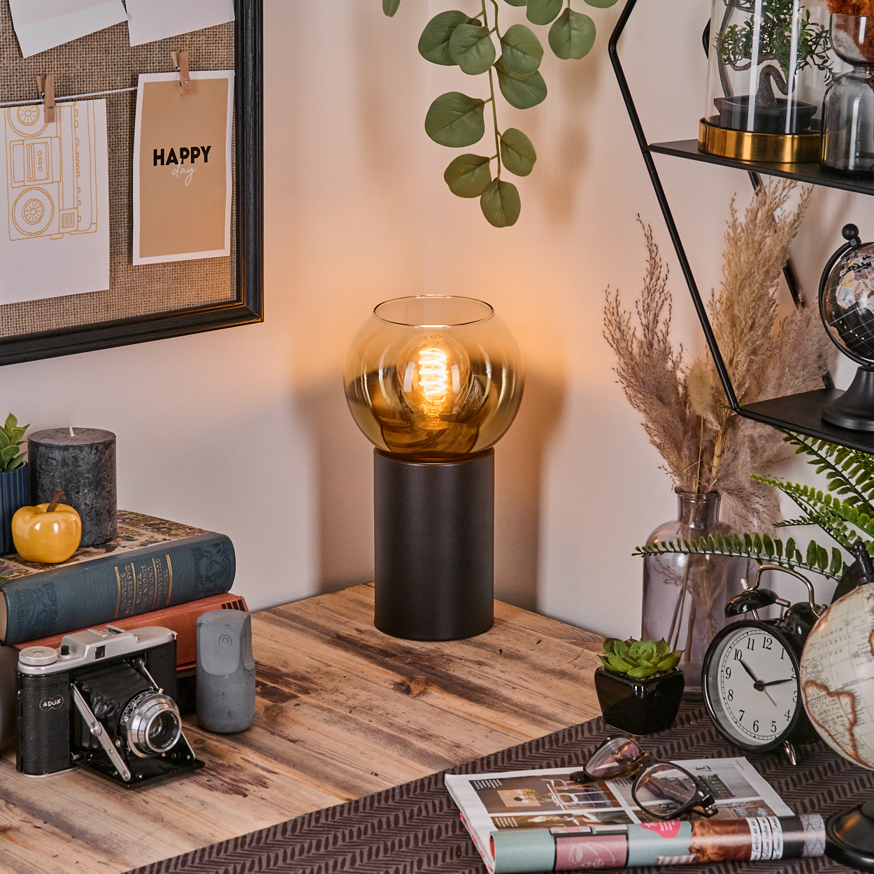 Ein Schreibtisch mit Lampe, Kamera, Büchern und Dekoration. Ein 'Happy Day'-Schild hängt an einer Tafel. Globus und Uhr rechts.