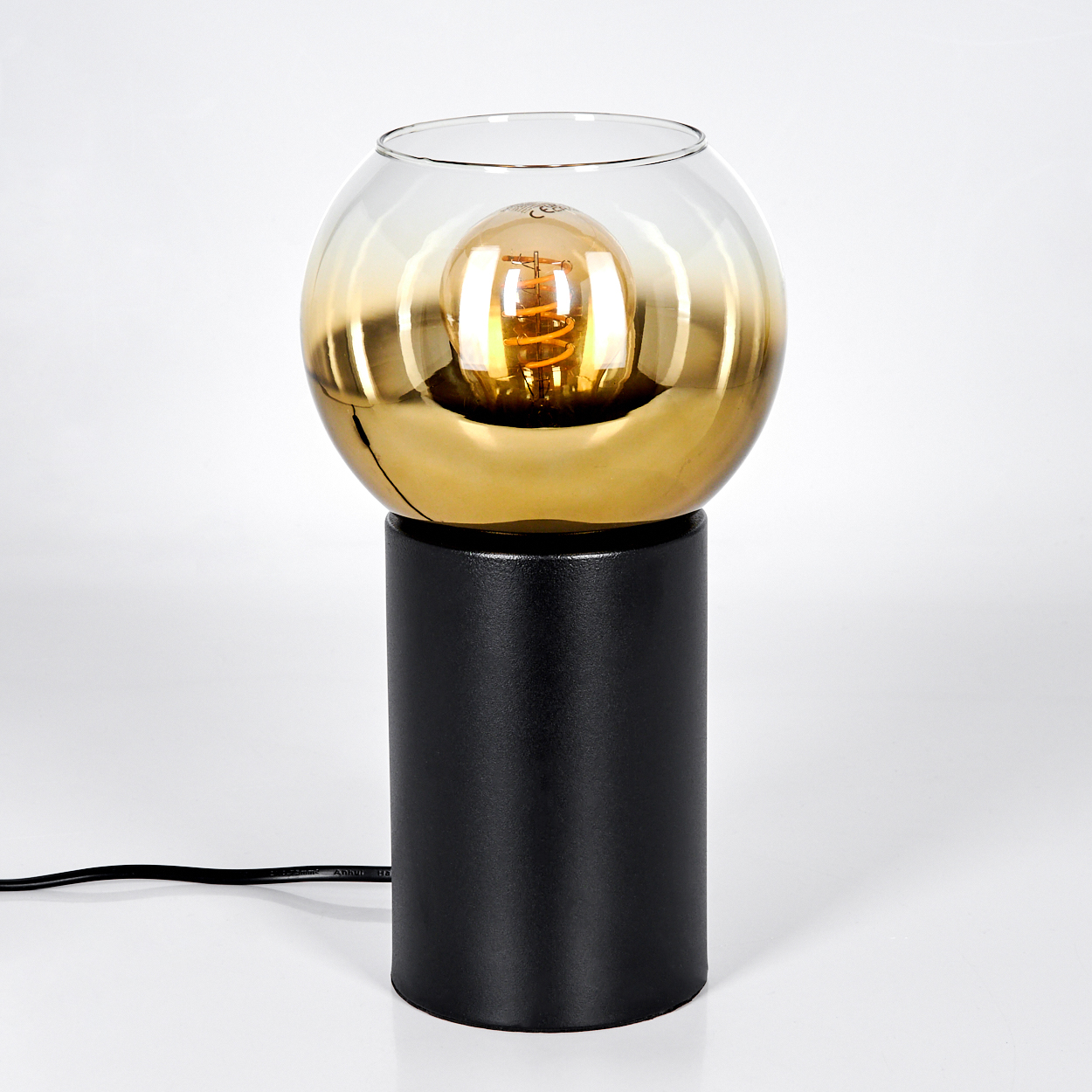 Eine moderne Lampe mit schwarzem Sockel und goldfarbenem und klarem Glasschirm, der eine Glühbirne enthält. Ein schwarzes Netzkabel ist sichtbar.