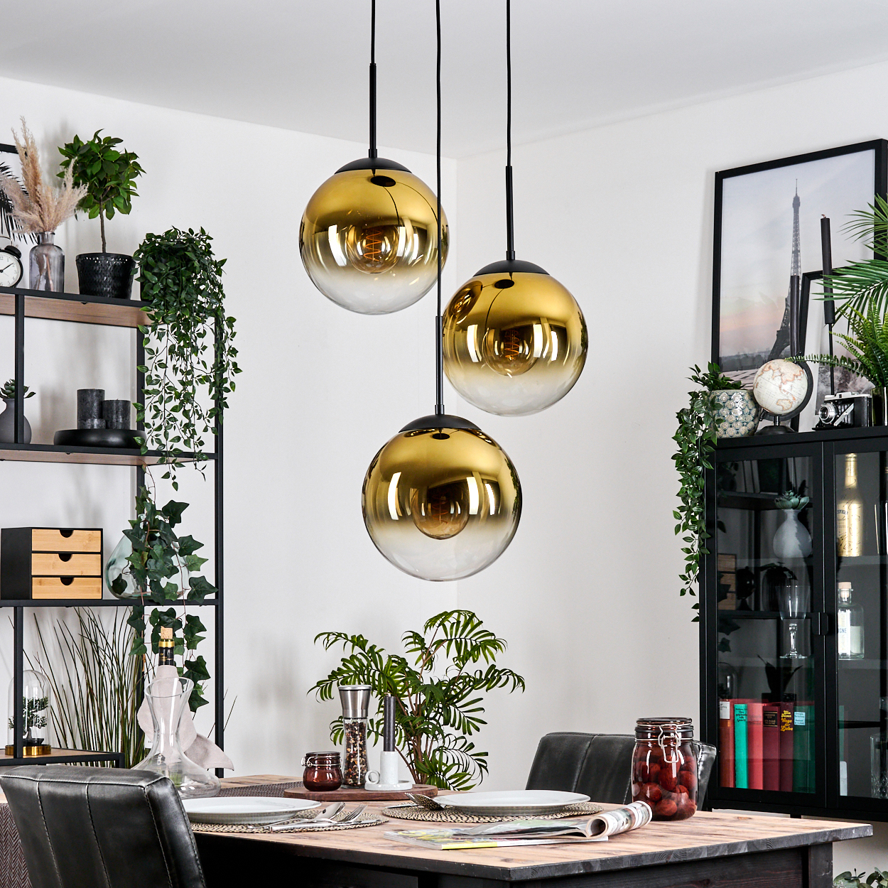 Esszimmer mit drei goldenen, kugelförmigen Pendelleuchten. Ein Tisch ist gedeckt. Regale und ein Schrank halten Pflanzen und Dekoration.