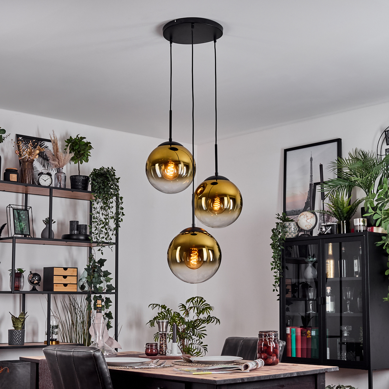 Drei goldfarbene Pendelleuchten hängen über einem Esstisch. Im Hintergrund Regale mit Pflanzen und Dekoration sowie ein Schrank.