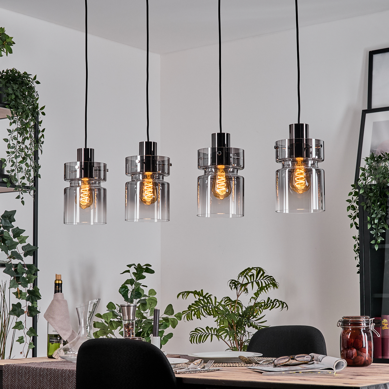 Vier Hängelampen mit sichtbaren Glühbirnen über einem Tisch, der für eine Mahlzeit gedeckt ist. Pflanzen und Dekoration umgeben den Essbereich.