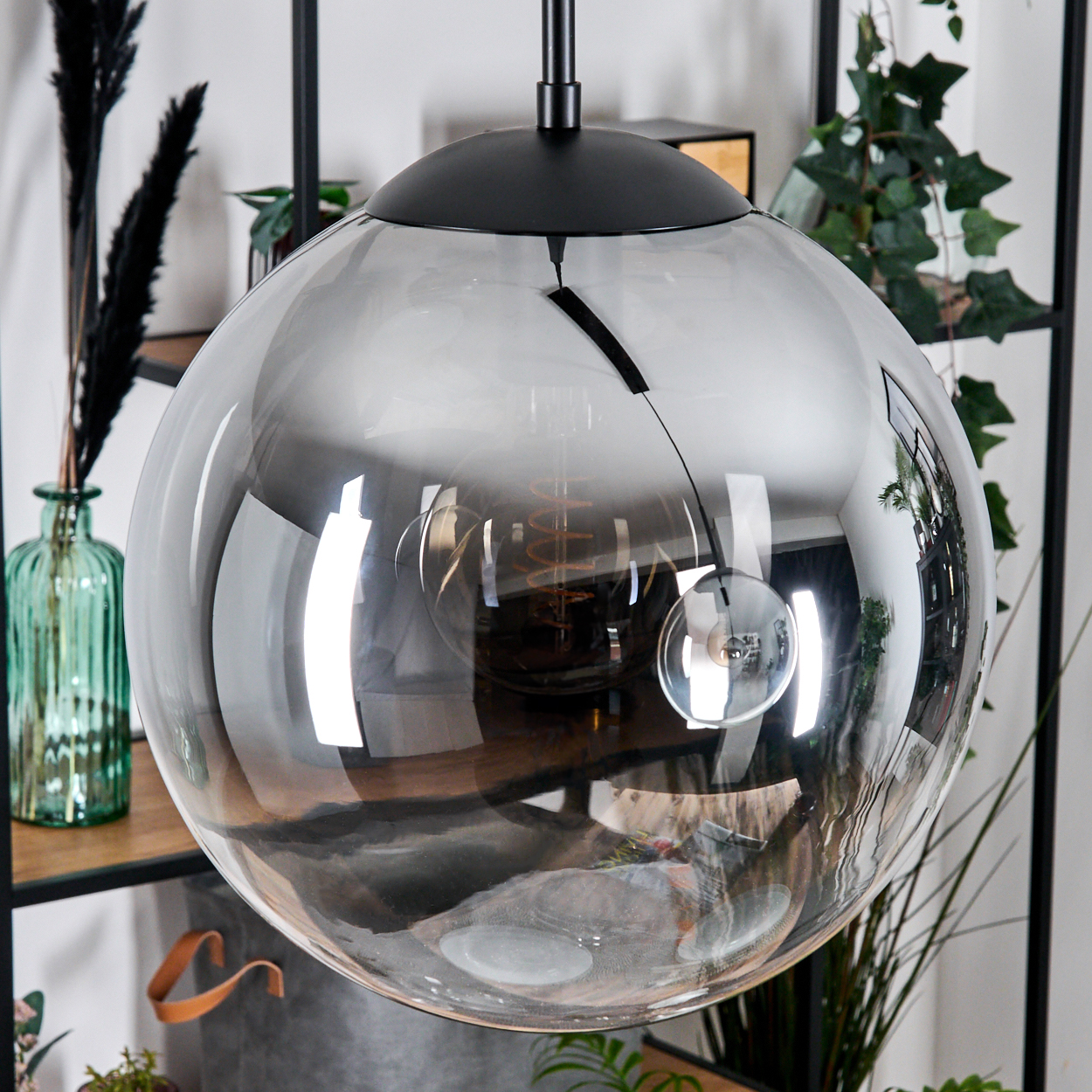 Eine große, graue Glaskugelleuchte hängt. Sie reflektiert einen Raum mit Pflanzen und einem Holzregal. Auf dem Regal steht eine grüne Vase.