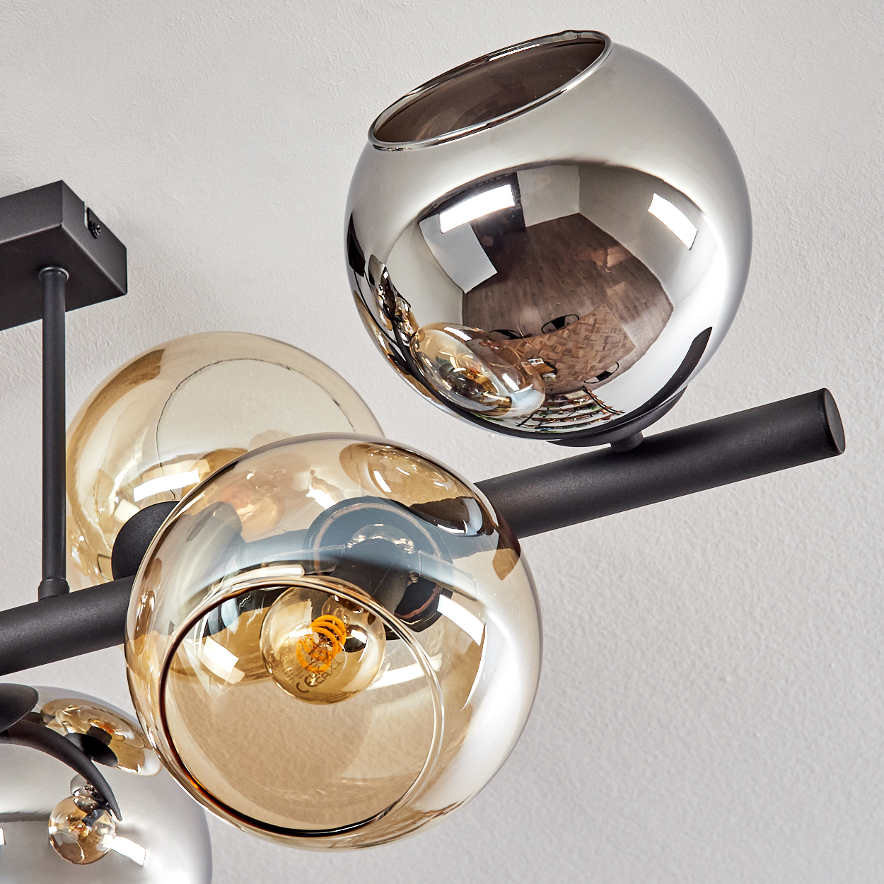 Eine moderne Deckenlampe mit Glaskugeln, einige bernsteinfarben und einige chromfarben. Ein schwarzer Metallrahmen hält die Kugeln. Weißer Wandhintergrund.