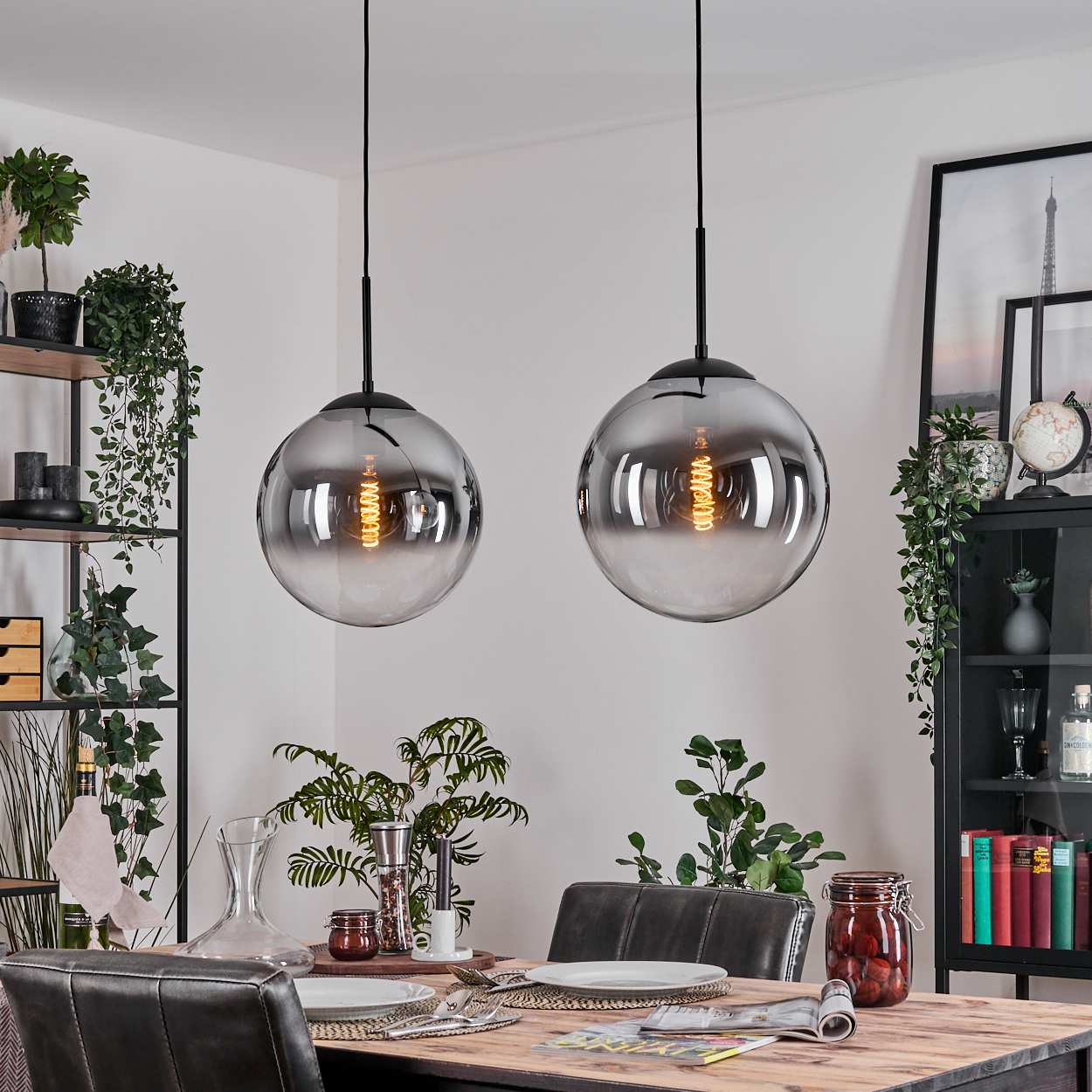 Zwei Kugellampen hängen über einem Esstisch mit Tellern und Besteck. Pflanzen und ein Schrank sind im Hintergrund.