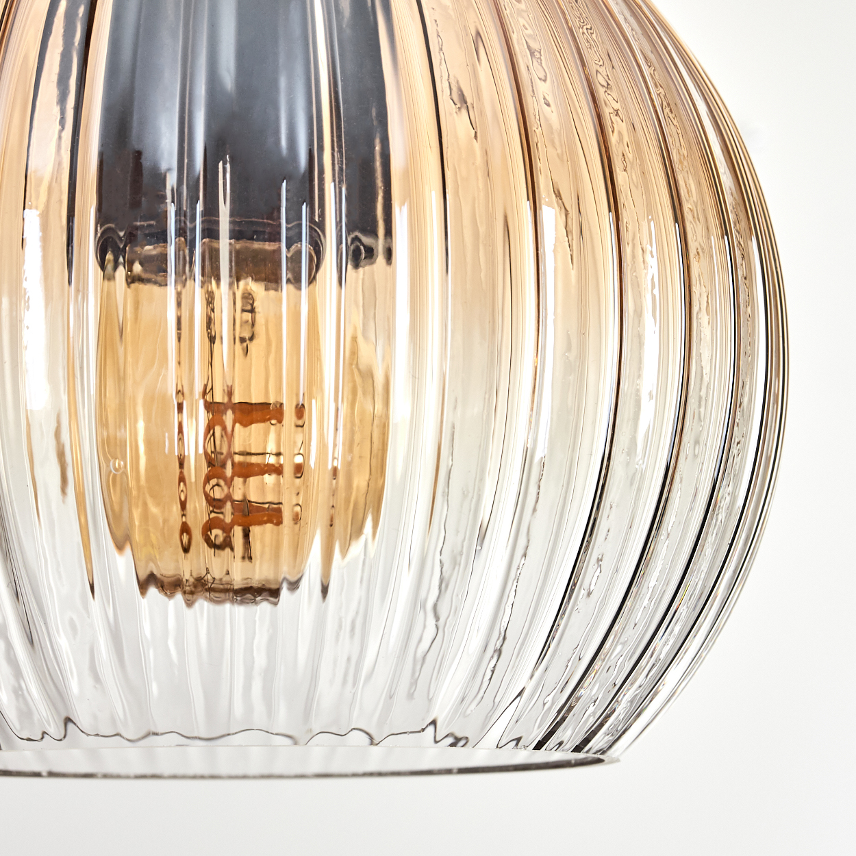 Nahaufnahme eines gerippten Glasschirms. Innen eine Glühbirne mit sichtbaren Filamenten. Hellbernsteinfarbenes und klares Glas vor weißem Hintergrund.