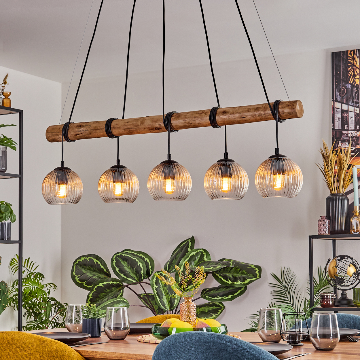 Ein Esszimmer mit Holztisch, Hängelampen und Pflanzen. Tisch mit Gläsern und Tellern gedeckt. Ein Gemälde an der Wand.