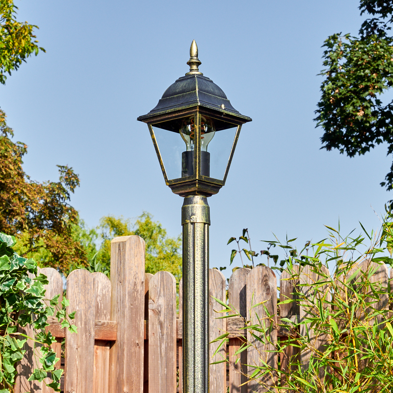 Eine dekorative Außenlampe mit Glasgehäuse steht vor einem Holzzaun, Grün und einem klaren blauen Himmel.