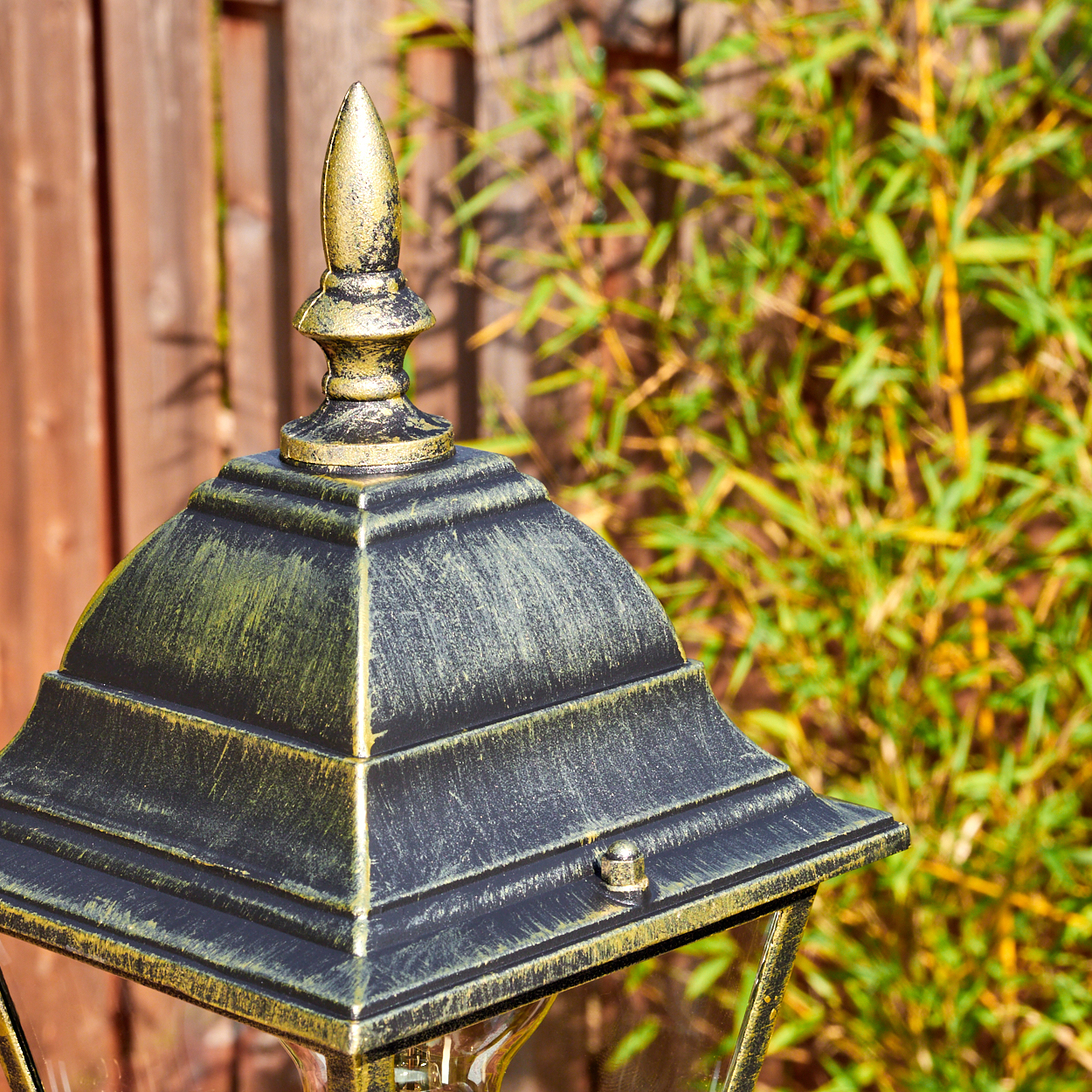 Nahaufnahme einer schwarz-goldenen Außenlampe. Ein Holzzaun und grüner Bambus im Hintergrund.