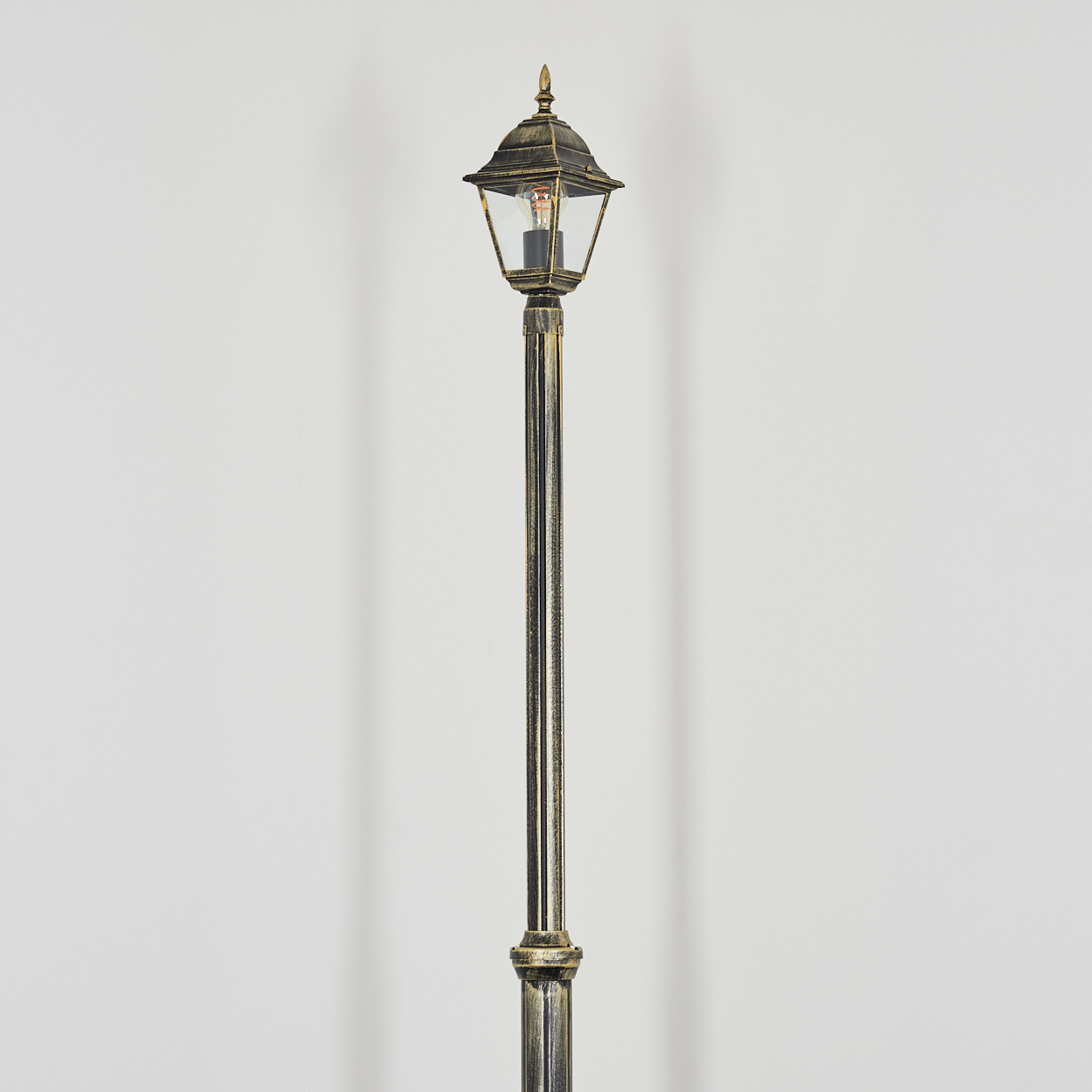 Eine verzierte, bronzefarbene Straßenlaterne steht vor einem schlichten weißen Hintergrund. Die Lampe ist beleuchtet.