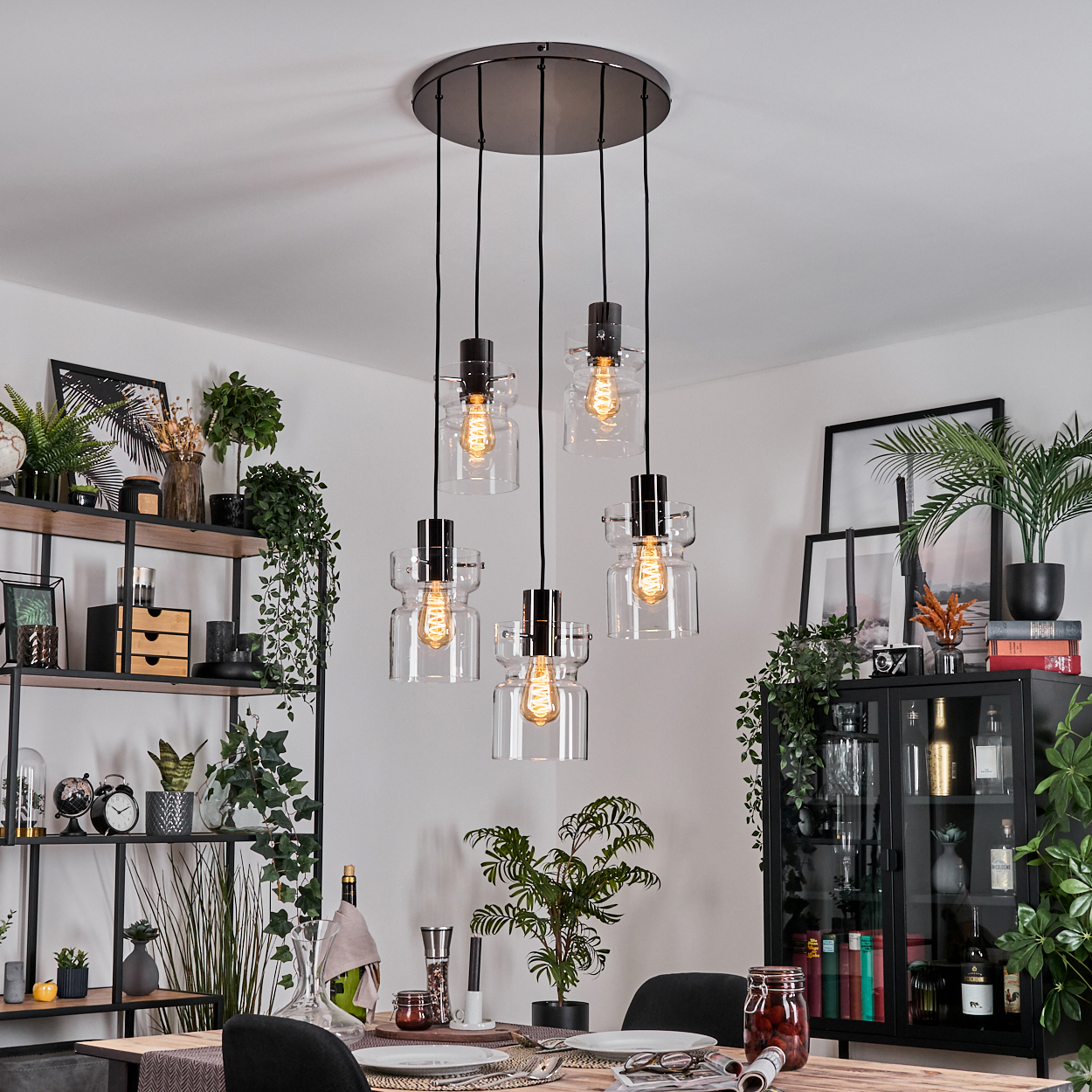 Esszimmer mit Hängelampen, Pflanzen und einem gedeckten Tisch. Ein schwarzer Schrank und Regale sind im Hintergrund.