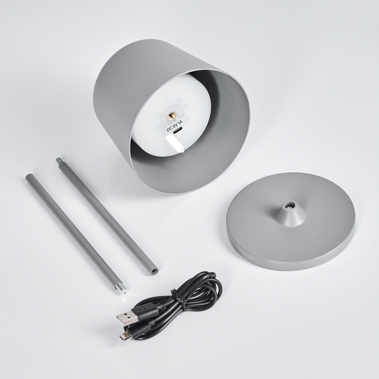 Eine zerlegte graue Lampe mit Teilen: zylindrischer Schirm, Sockel, zwei Stäbe, ein USB-Kabel. Der Schirm zeigt eine weiße Lichtkomponente.