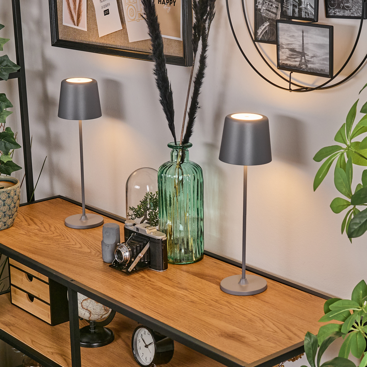 Zwei graue Lampen, eine grüne Vase und eine alte Kamera stehen auf einem Holzregal. Eine Uhr und ein Globus befinden sich darunter.