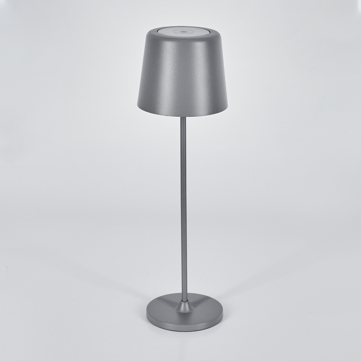 Eine graue Tischlampe steht auf einer weißen Oberfläche. Sie hat einen Lampenschirm, einen langen Stab und einen runden Fuß. Der Hintergrund ist weiß.