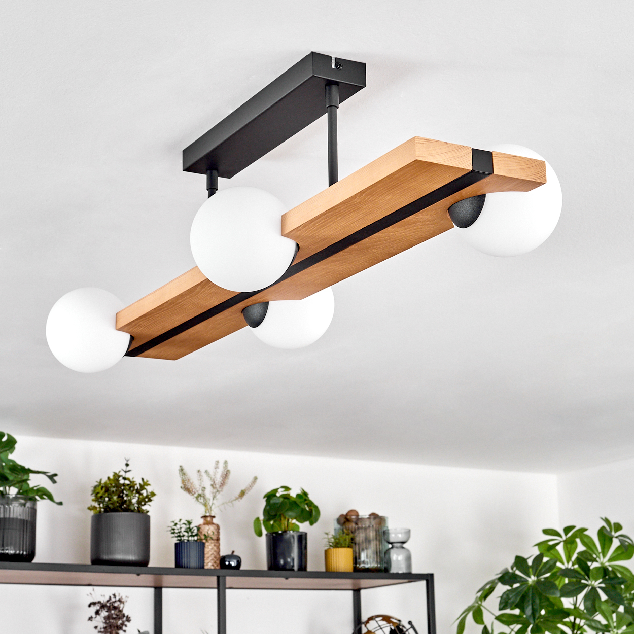 Eine moderne Deckenlampe mit vier weißen Kugeln. Sie hat einen Holzbalken mit schwarzen Akzenten und einen schwarzen Metallsockel. Pflanzen sind unten sichtbar.