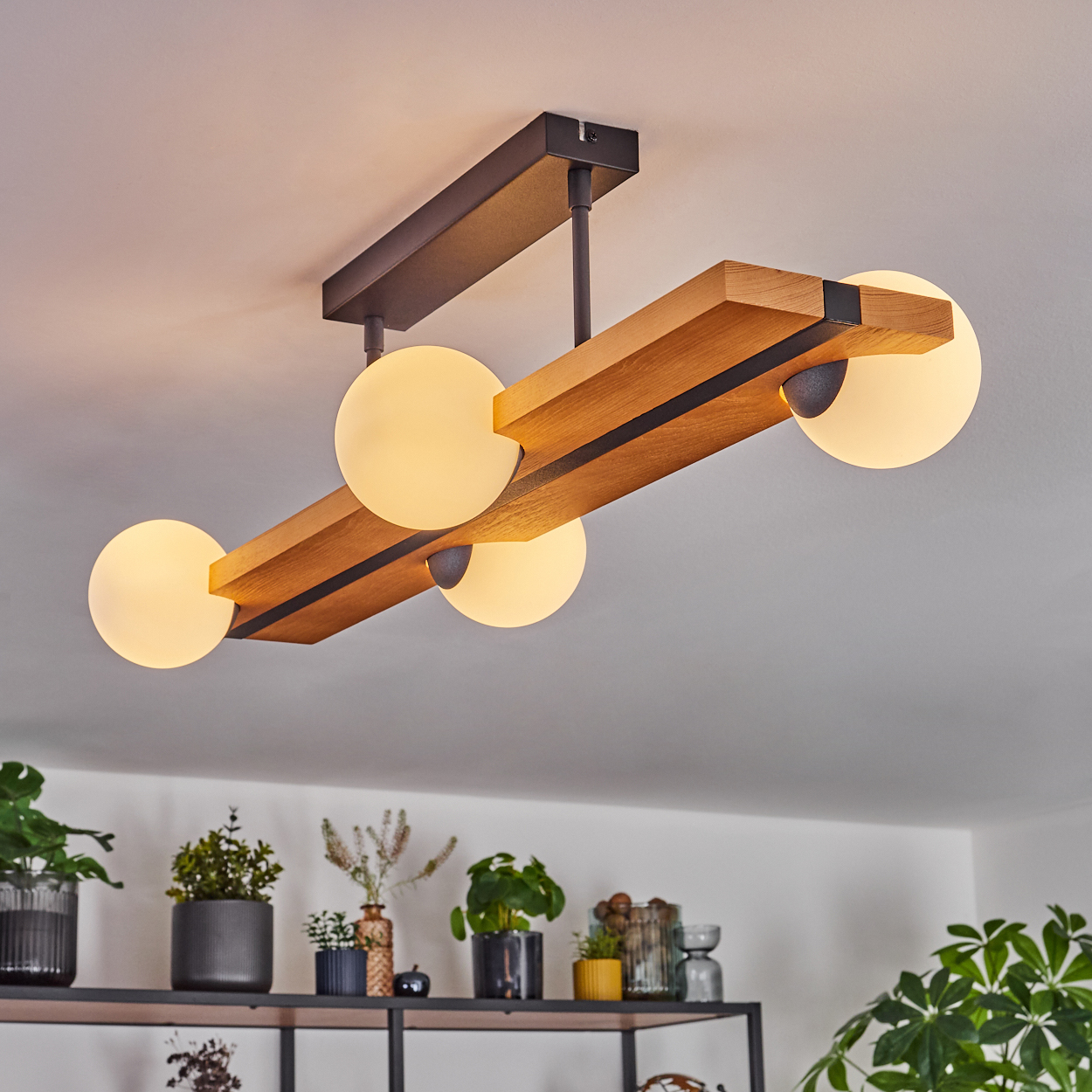 Eine moderne Deckenlampe mit vier leuchtenden Kugeln. Die Lampe besteht aus Holz und schwarzem Metall. Pflanzen und Regale sind im Hintergrund.