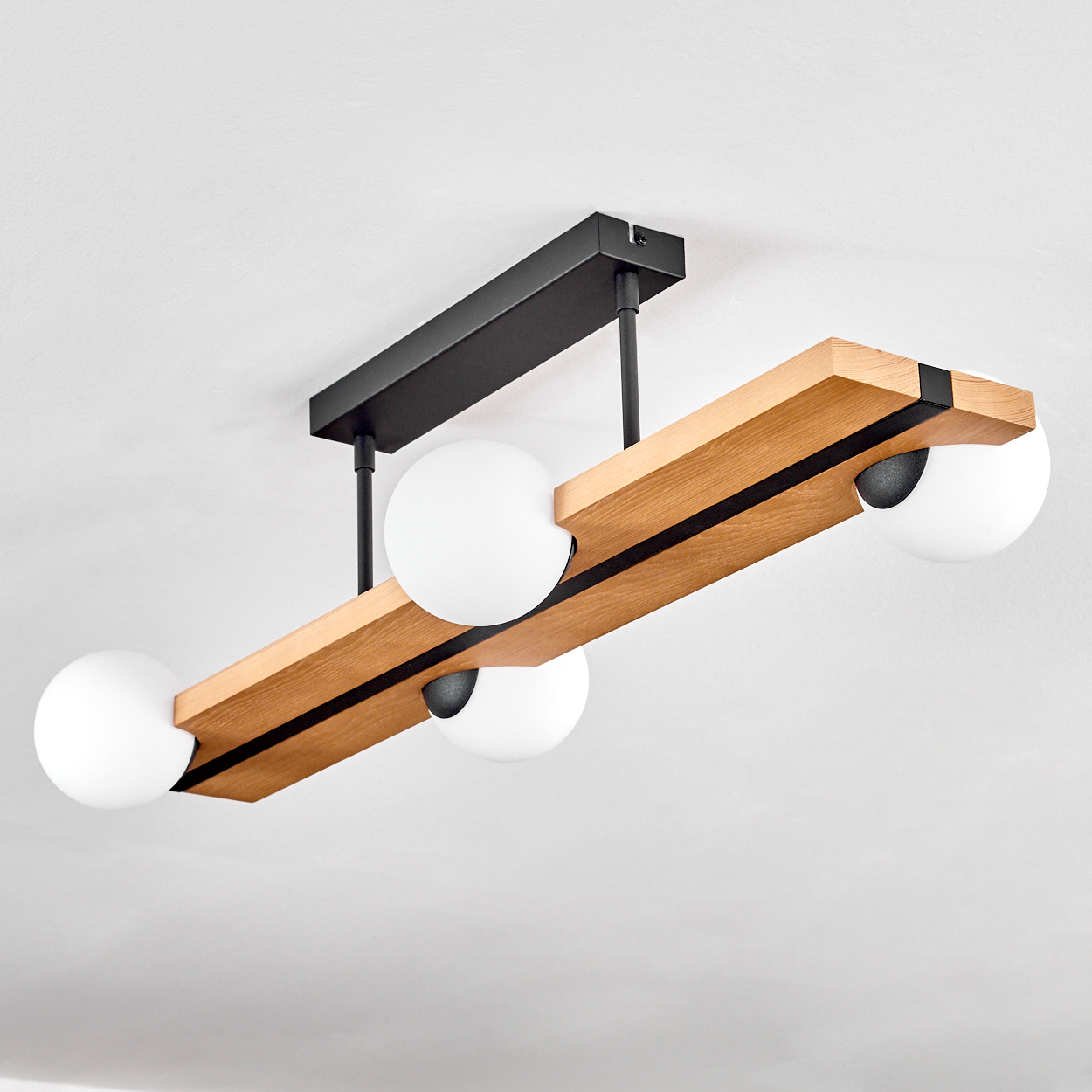 Eine moderne Deckenlampe mit Holzbalken, schwarzen Metallakzenten und vier weißen Kugelleuchten. Der Hintergrund ist eine weiße Decke.