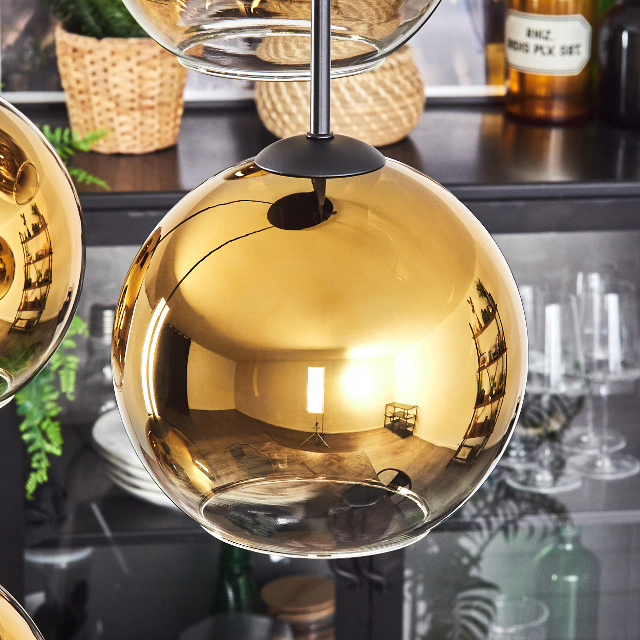 Eine goldene, kugelförmige Pendelleuchte spiegelt einen Raum wider. Im Hintergrund sind Pflanzen in Körben und ein Regal mit Flaschen zu sehen.