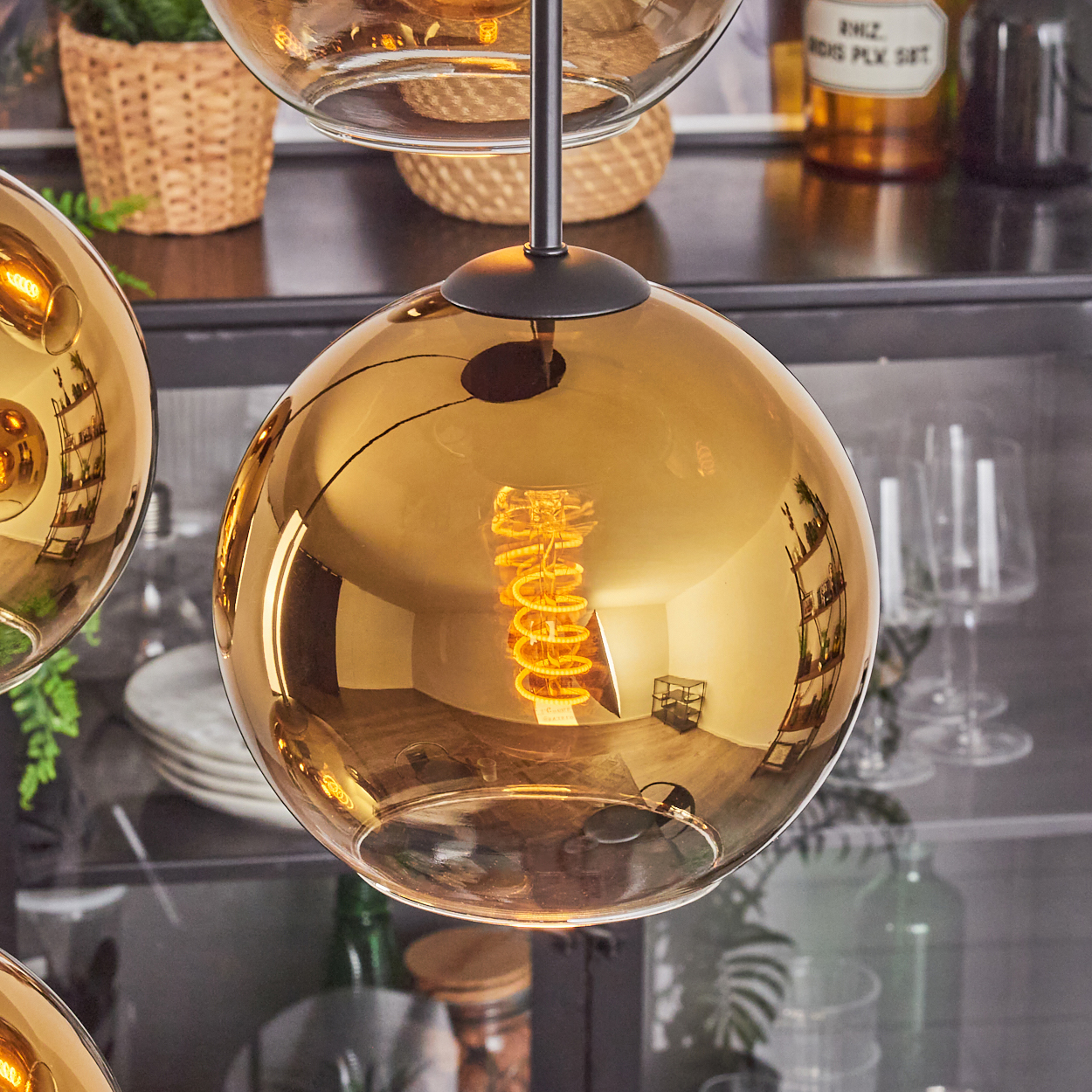 Nahaufnahme einer goldenen Glas-Pendelleuchte mit sichtbarem Wendel-Glühfaden. Hintergrund ist ein Regal und andere Lampen.