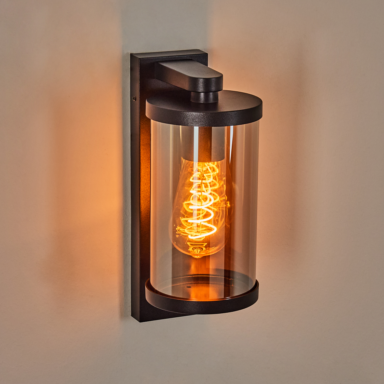 Schwarze Wandleuchte mit Glaszylinder. Im Inneren befindet sich eine leuchtende Wendelglühbirne. Beigefarbene Wand im Hintergrund.