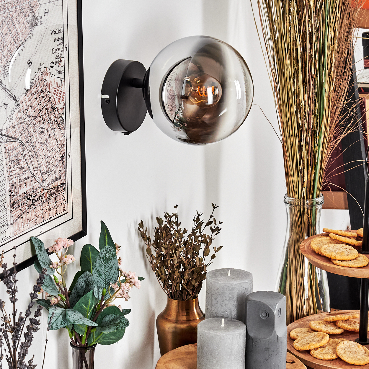 Eine moderne Wandlampe mit kugelförmigem Glasschirm, Karte, Blumen, Kerzen und Keksen auf einem Holztablett.