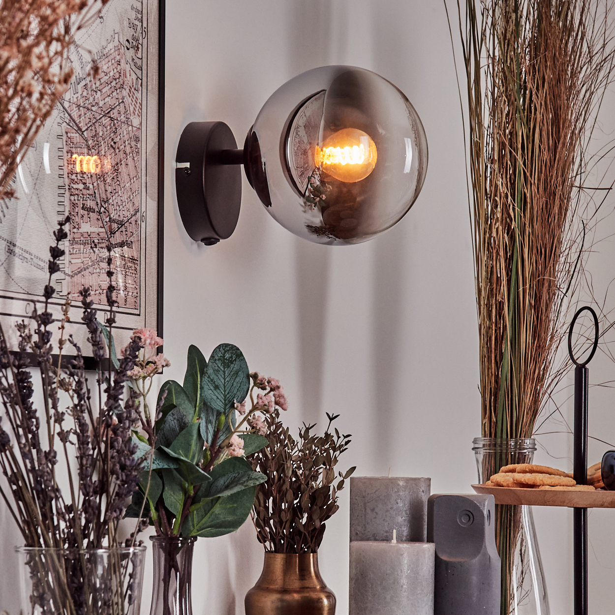Eine Wandlampe mit Globus-Schirm, leuchtende Glühbirne. Dekorationen umfassen Blumen, Kerzen und einen Kartendruck.