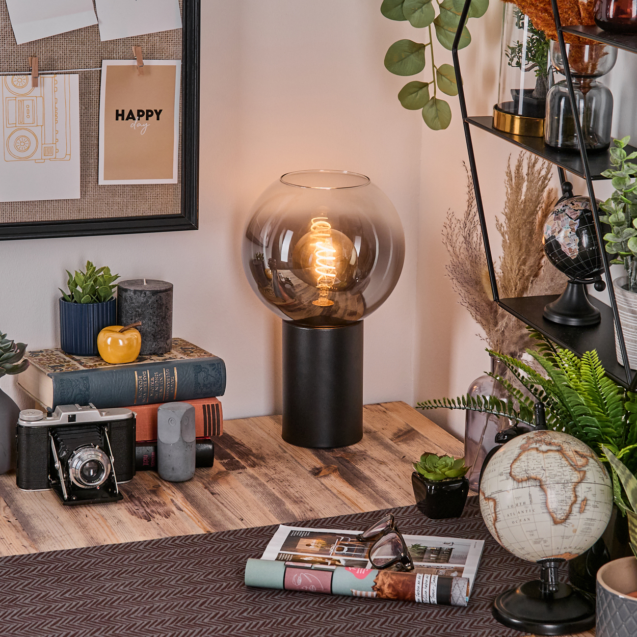 Ein Schreibtisch mit Lampe, Büchern, Kamera, Globus und Pflanzen. Eine 'Happy'-Notiz im Hintergrund.