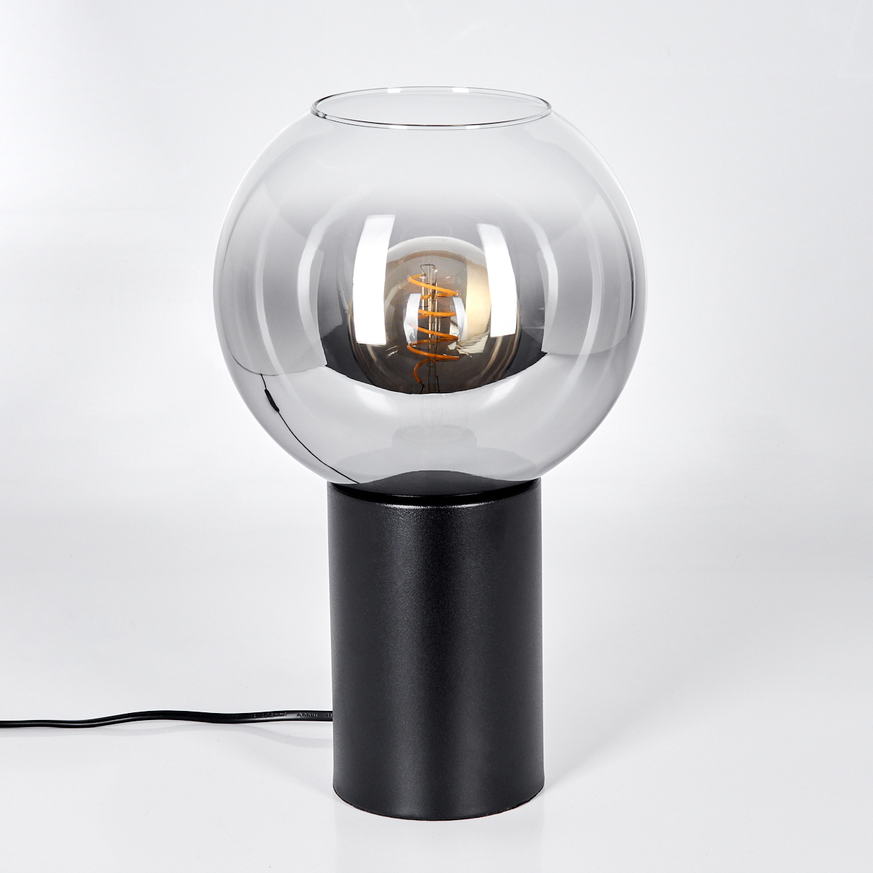Eine moderne Lampe mit schwarzem Sockel und rundem, rauchigem Glasschirm. Eine Glühbirne ist im Schirm zu sehen. Ein schwarzes Kabel ist sichtbar.
