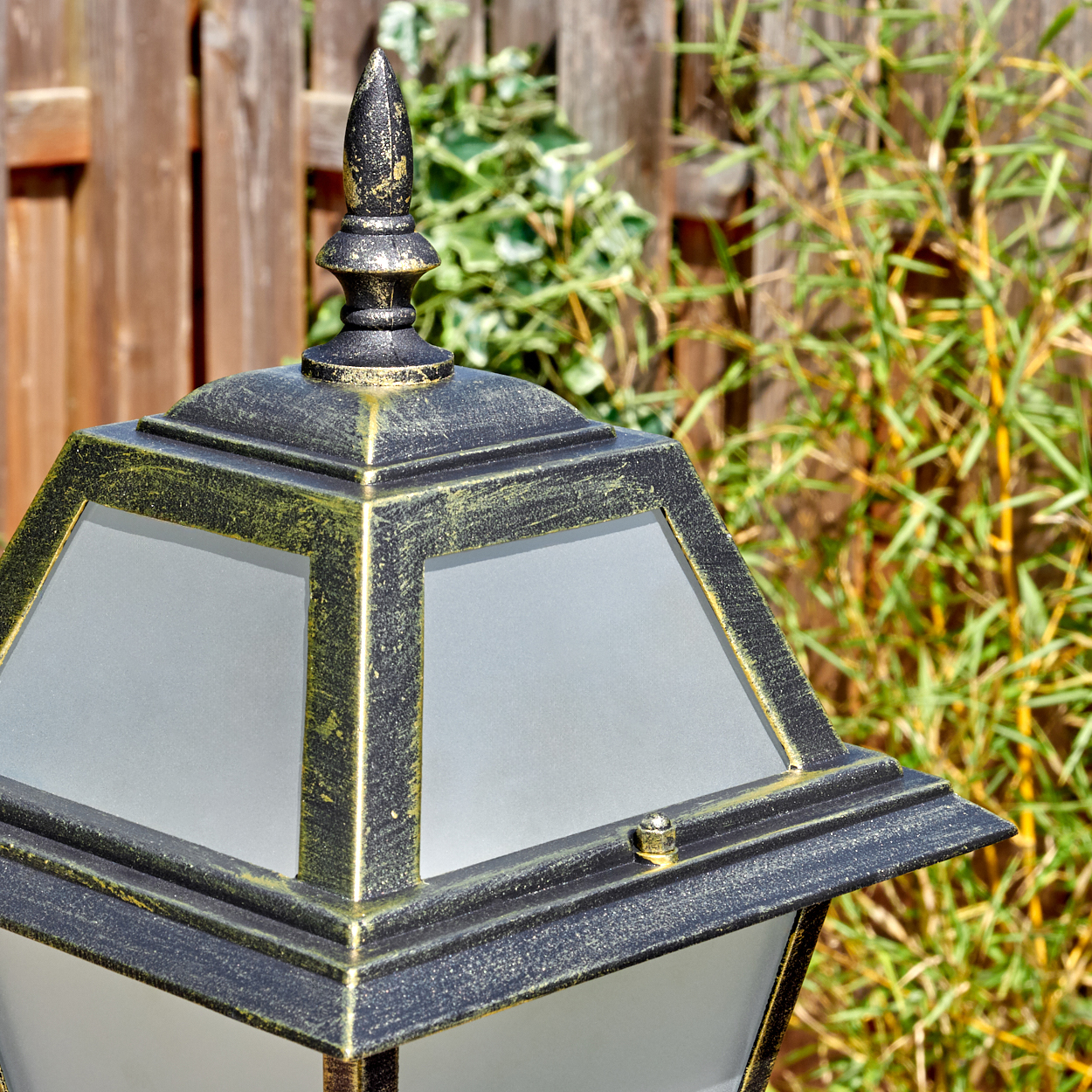 Nahaufnahme einer schwarzen und goldenen Außenlampe. Ein Holzzaun und grüne Pflanzen befinden sich im Hintergrund.