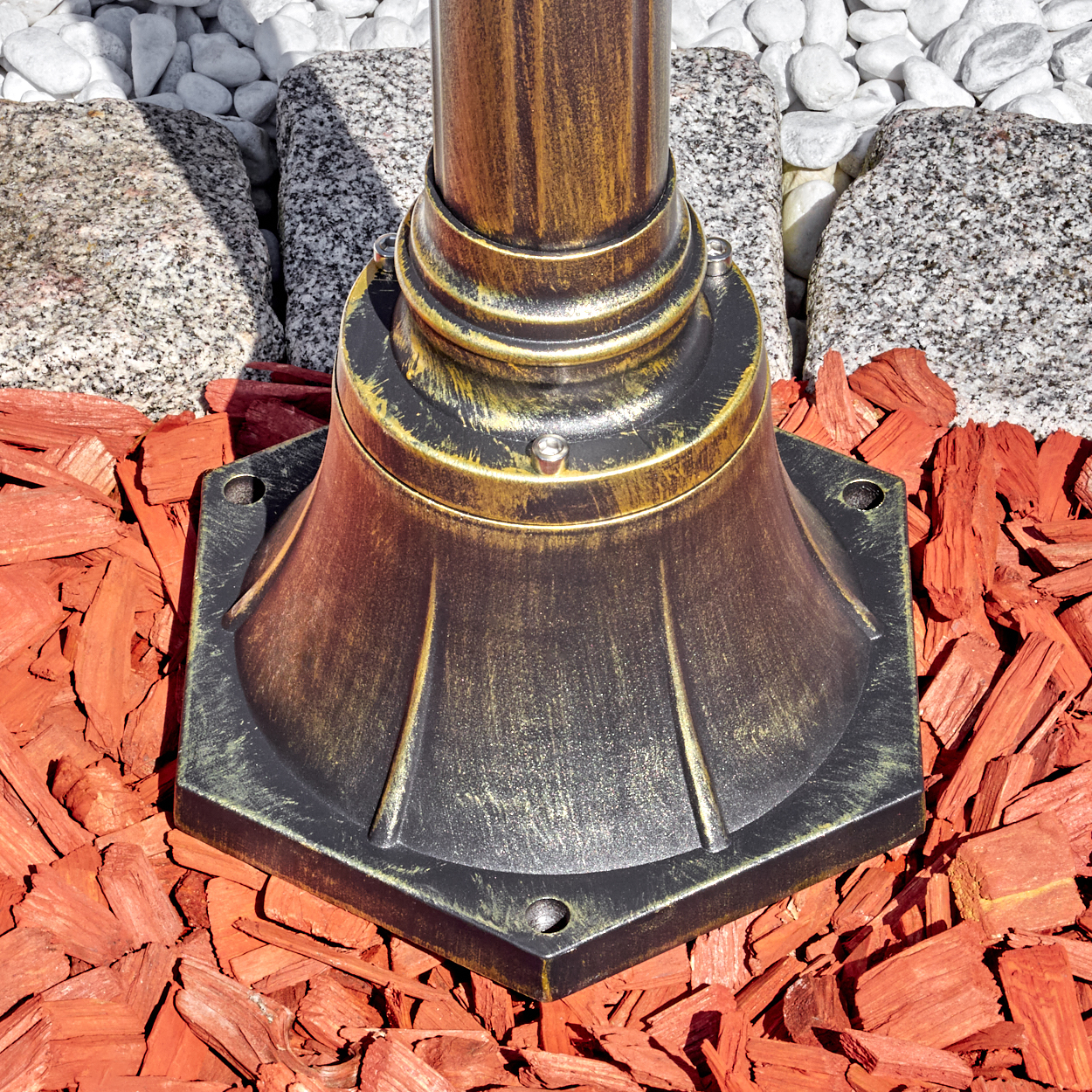Ein bronzefarbener Lampensockel auf roter Mulch, mit grauem Granit und weißen Steinen im Hintergrund. Die Lampe ist verziert und gealtert.
