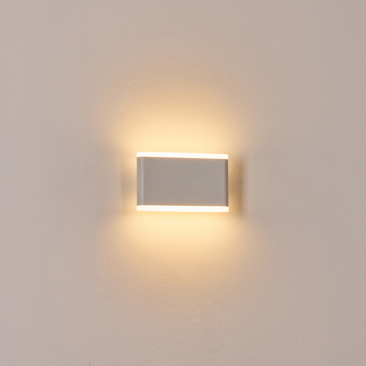 Eine rechteckige, graue Wandlampe leuchtet. Sie ist an einer hellbeige Wand montiert. Das Licht scheint von oben und unten.