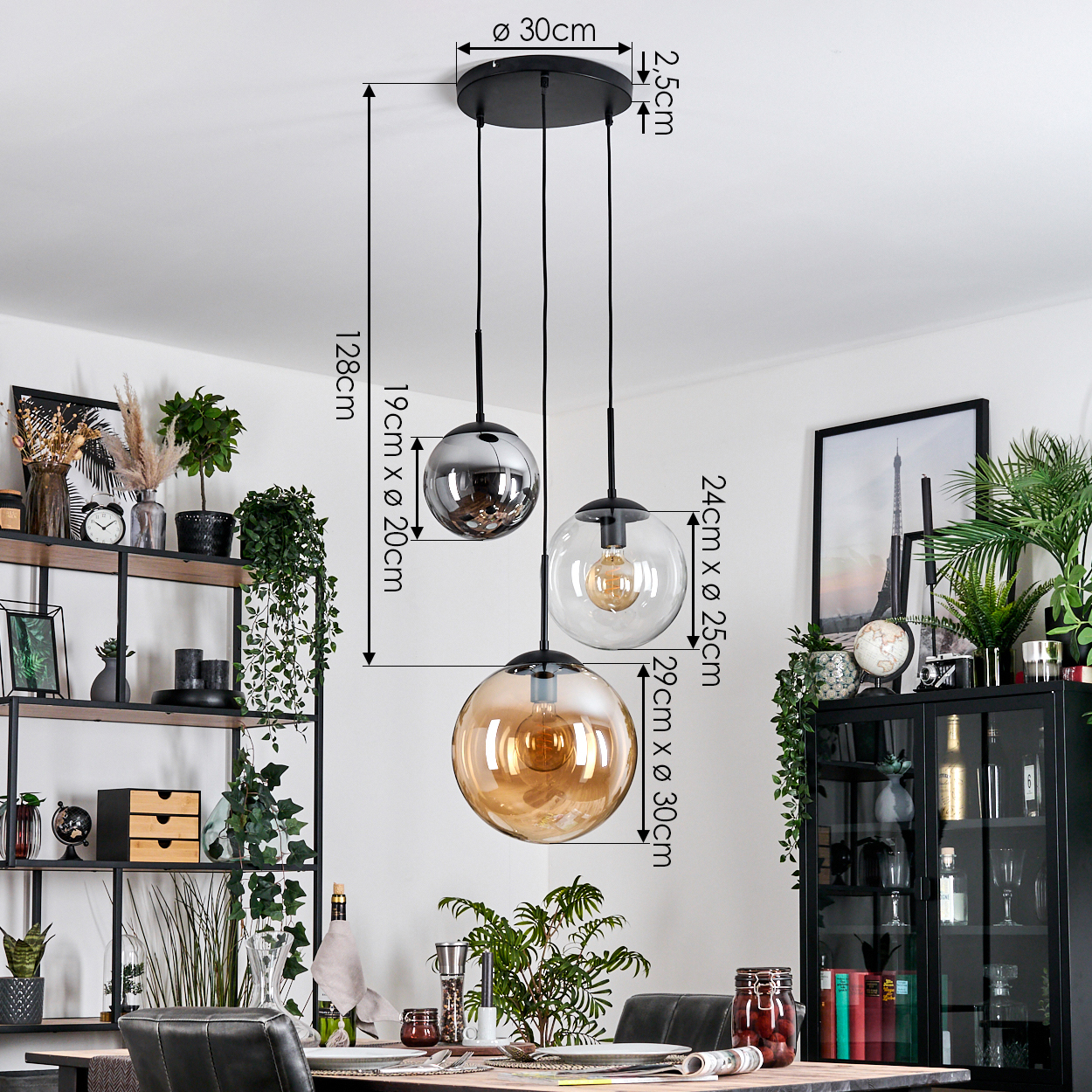 Drei Pendelleuchten mit Glaskugeln hängen von einer schwarzen Deckenleuchte. Das Zimmer hat Pflanzen, Möbel und ein Bild.