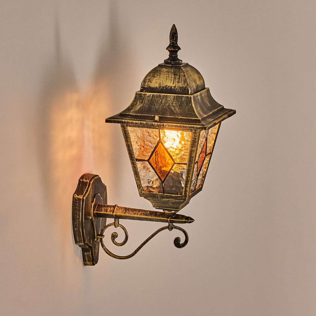 Eine verzierte, goldene Außenlampe an einer Wand. Die Lampe hat Buntglasscheiben, die einen warmen Schein und Schatten werfen.