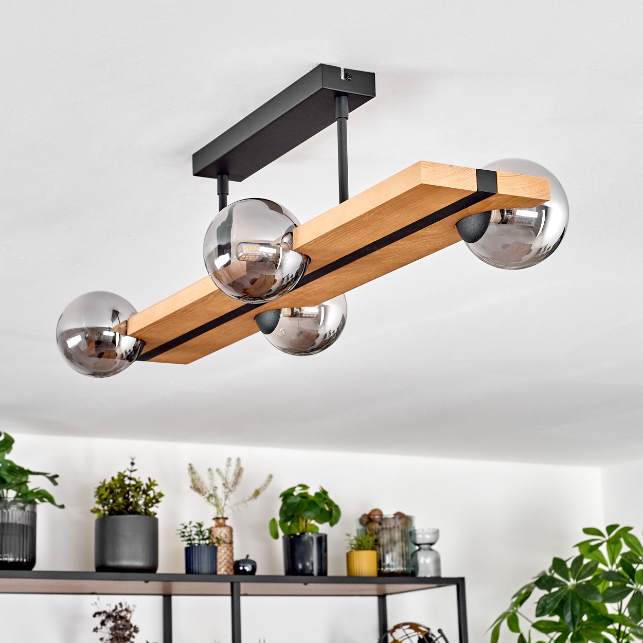Eine moderne Deckenlampe mit vier silbernen Kugeln. Sie besteht aus Holz und schwarzem Metall, darunter Pflanzen auf einem Regal.