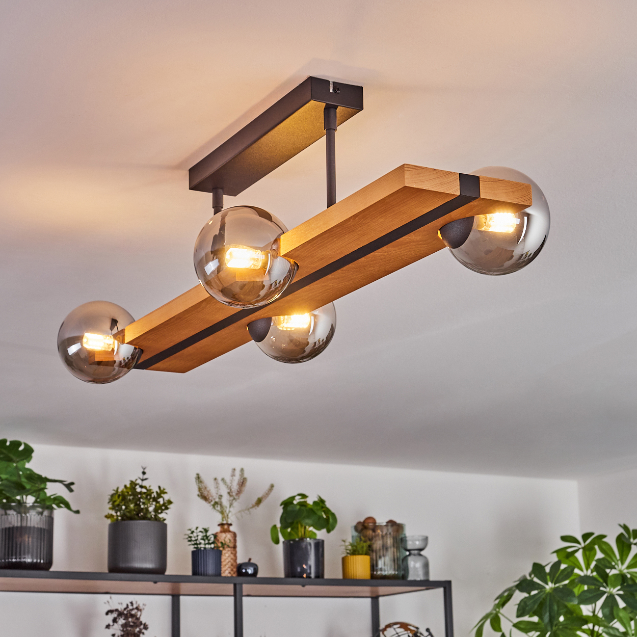 Eine moderne Deckenleuchte mit vier Glaskugeln hängt über einem Regal mit Pflanzen und Dekoration. Das Licht hat einen Holzbalken.