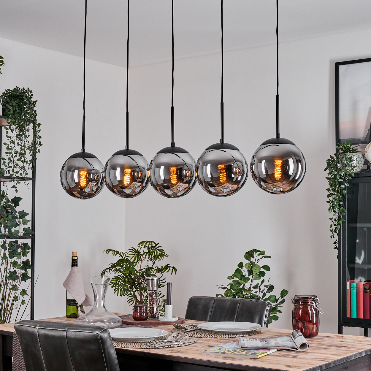 Esszimmer mit Holztisch, fünf Hängelampen, Pflanzen und einem schwarz gerahmten Bild an einer weißen Wand.