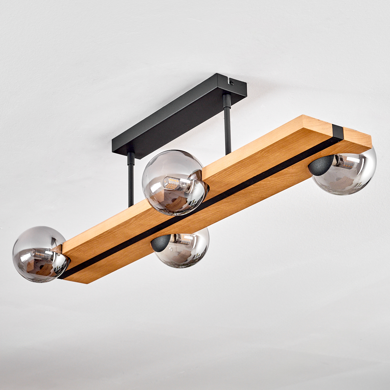 Eine moderne Deckenlampe. Sie hat einen Holzbalken mit vier kugelförmigen, reflektierenden Glaslampen. Die Halterung ist schwarz und an einer weißen Decke befestigt.