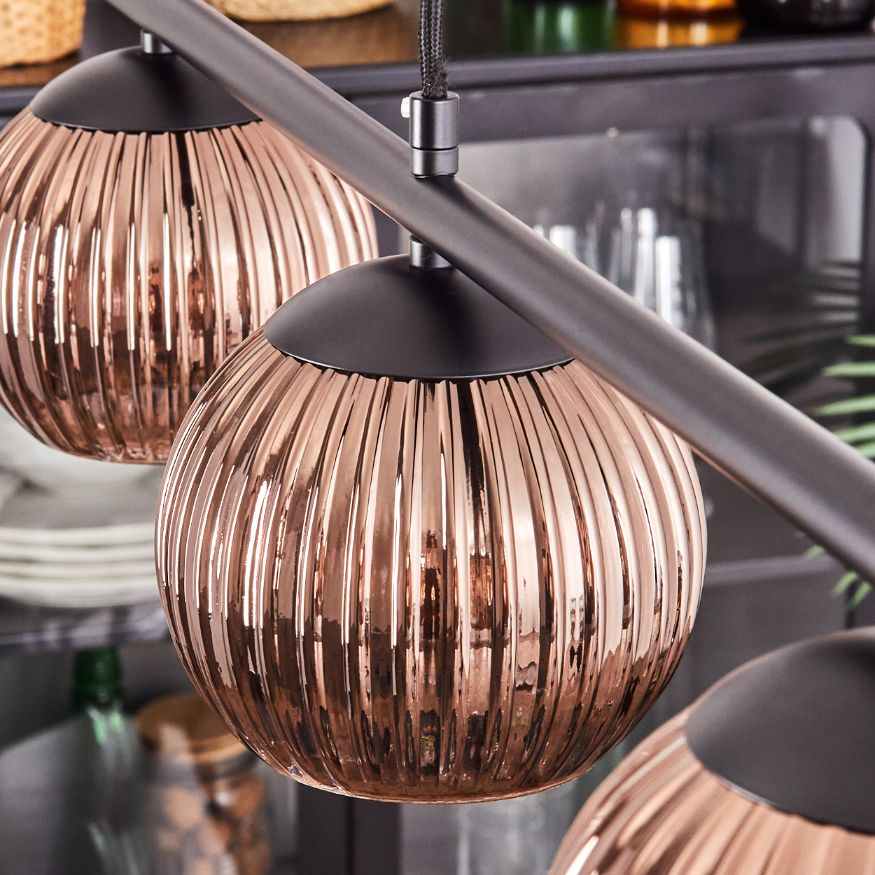 Nahaufnahme von drei kupferfarbenen, gerippten Glas-Pendelleuchten. Sie hängen an einer dunklen Metallstange. Hintergrund mit verschwommenen Küchenartikeln.