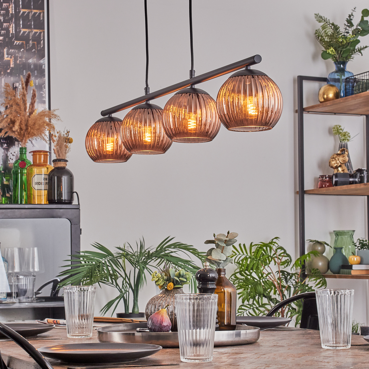 Esszimmer mit einem Holztisch, der zum Abendessen gedeckt ist. Darüber ist eine Lampe mit vier Glaskugeln zu sehen. Pflanzen und Dekoration sind im Hintergrund.