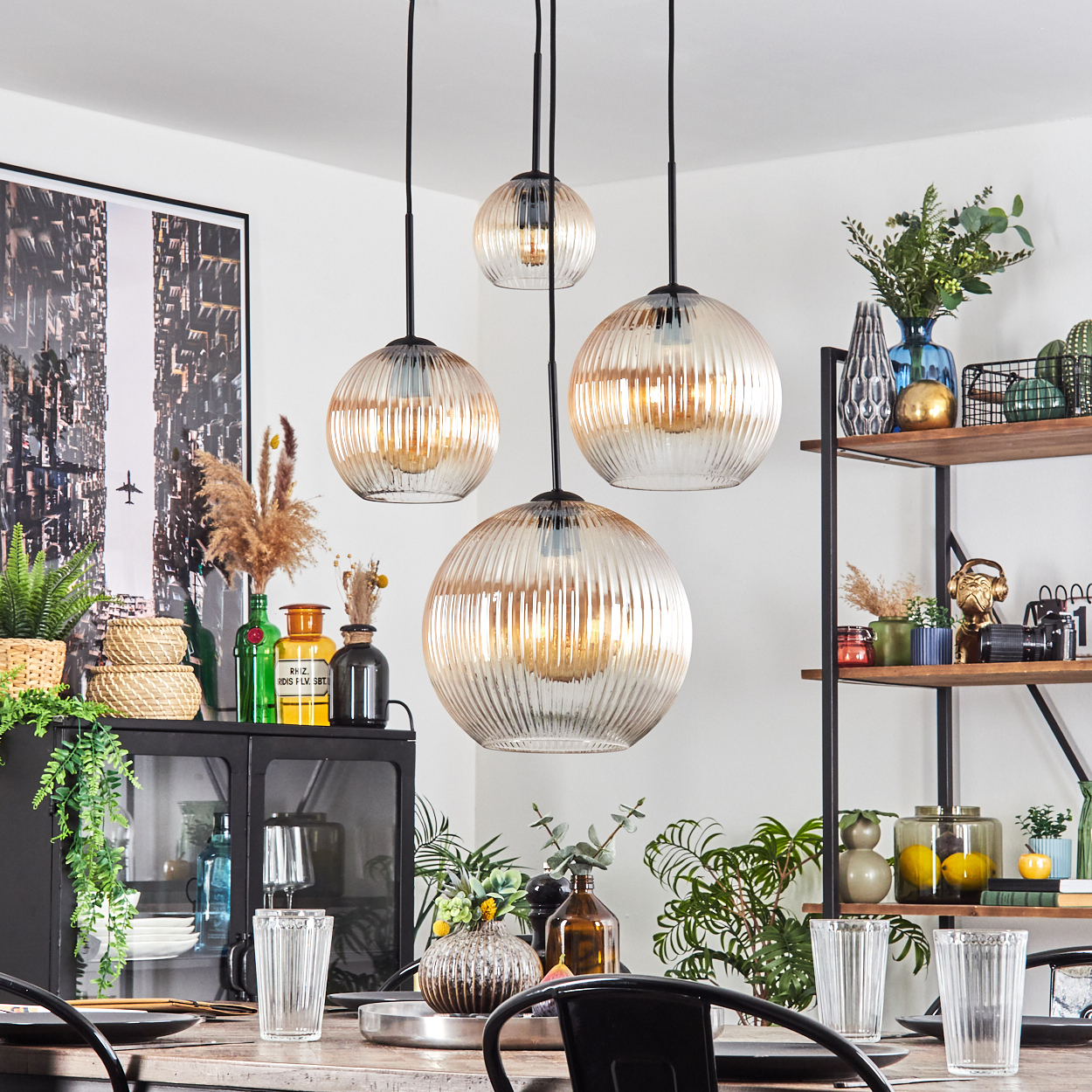 Esszimmer mit hängenden Kugelleuchten. Tisch mit Gläsern gedeckt. Pflanzen und Dekoration auf Regalen und Schrank. Kunstwerk.