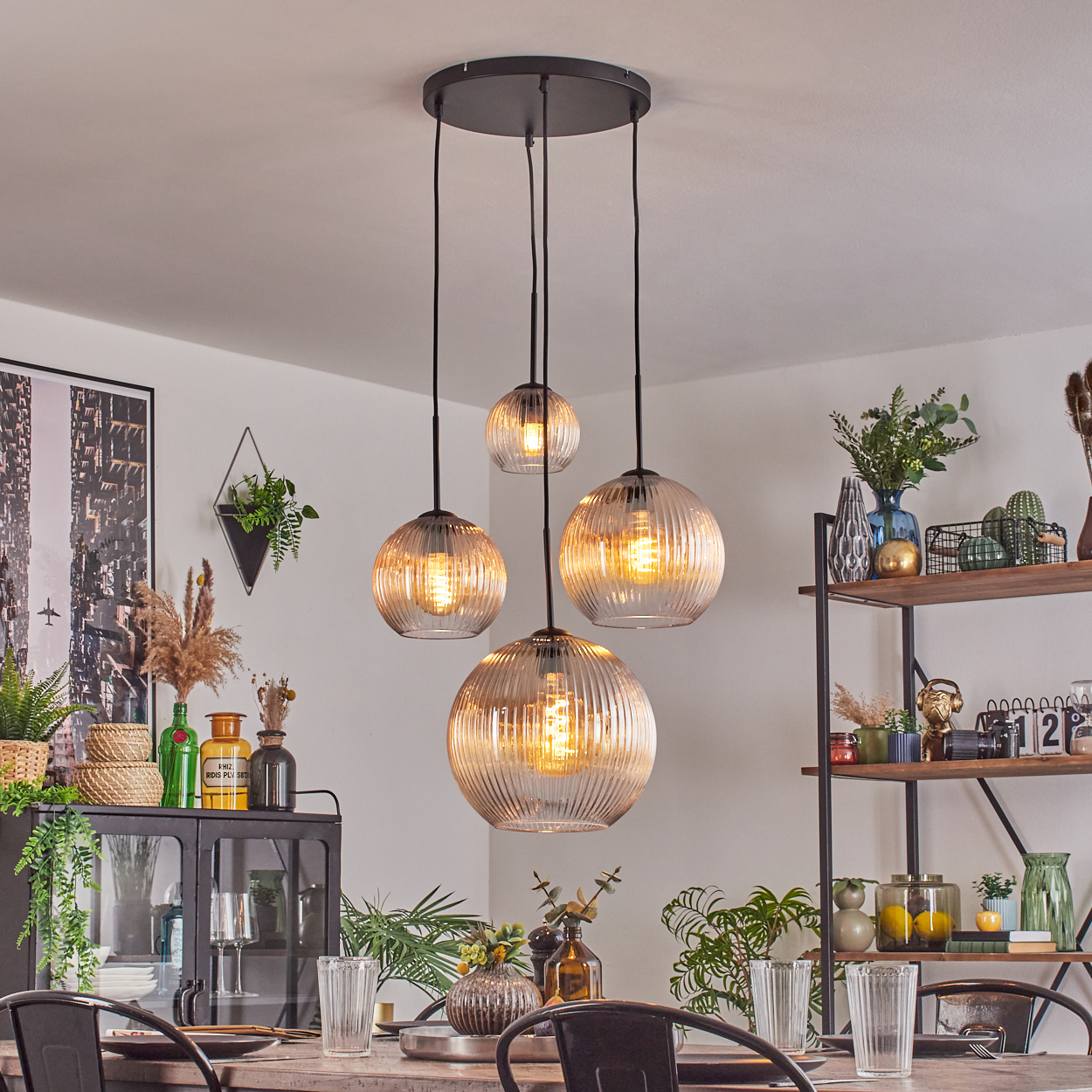 Esszimmer mit hängenden Kugelleuchten. Tisch mit Gläsern. Regale mit Pflanzen und Dekoration. Schwarzer Metallschrank mit Dekorationen.