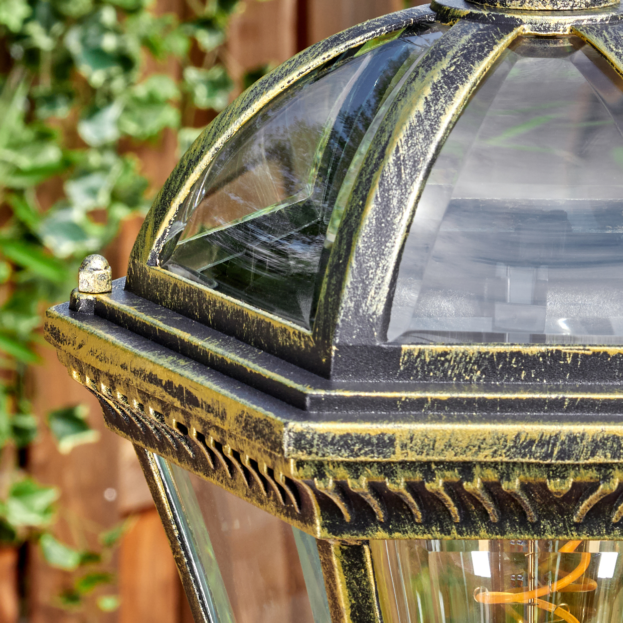 Nahaufnahme einer schwarz-goldenen Außenlampe. Glasscheiben spiegeln die Umgebung wider. Grünes Laub und Holzzaun im Hintergrund.