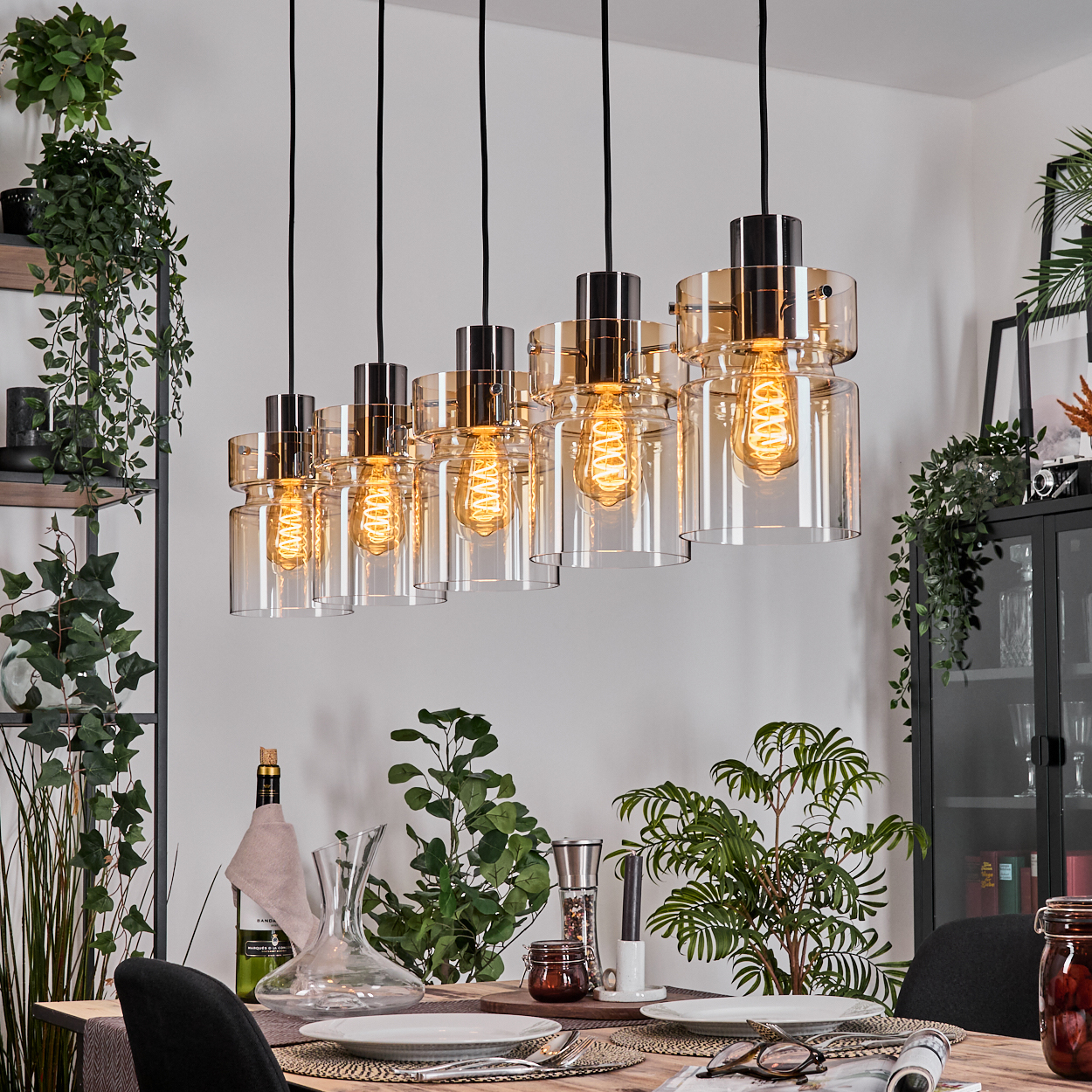Esszimmer mit Holztisch, Hängelampen, Weinflasche und Pflanzen. Eine Tischdecke mit Tellern und Gläsern.