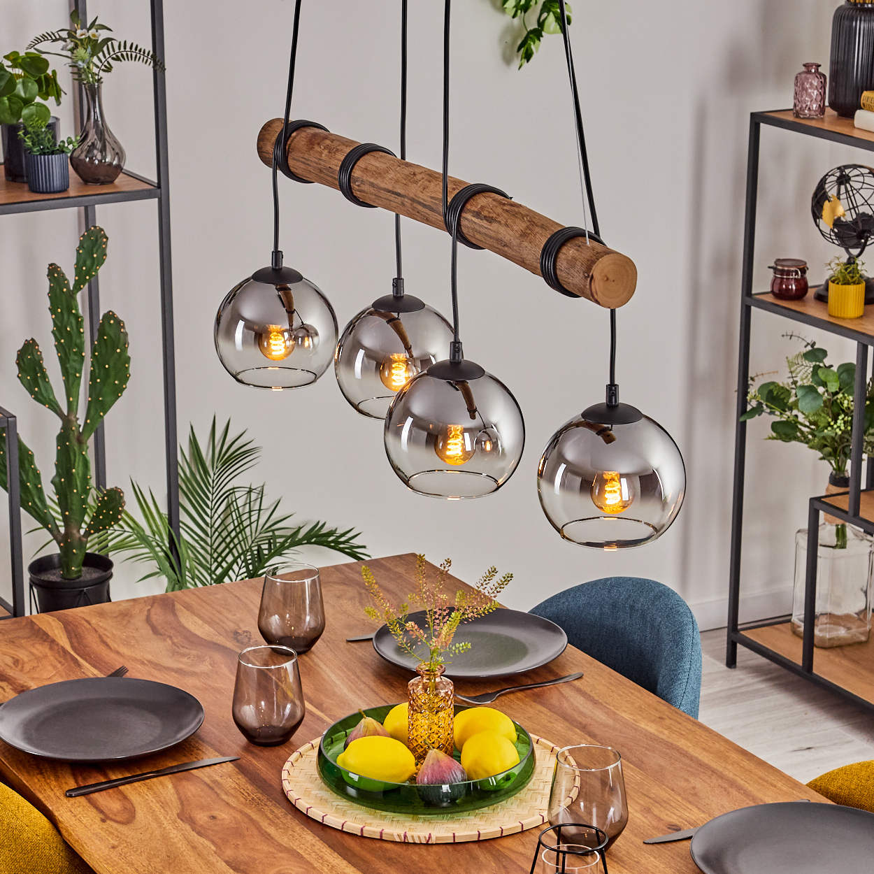 Ein Esstisch mit Holzplatte, gedeckt mit Tellern, Gläsern und einem Mittelstück. Darüber ein Holzbalken mit vier Hängelampen.