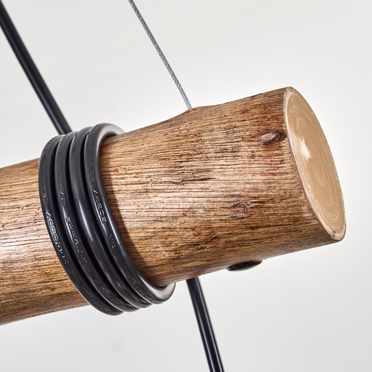 Nahaufnahme eines Holzstamms mit rundem Querschnitt, umwickelt mit schwarzen Kabeln und an einem Stahlseil aufgehängt.