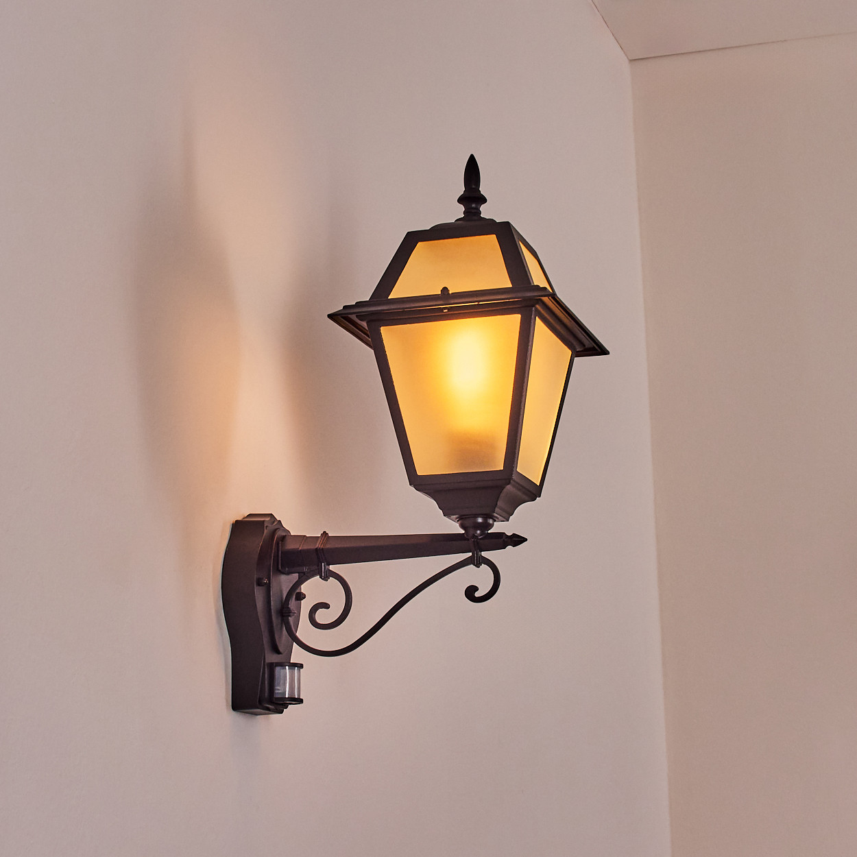 Dunkle Metall-Außenlampe an heller Wand. Die Lampe leuchtet warm. Ein Bewegungssensor ist sichtbar.