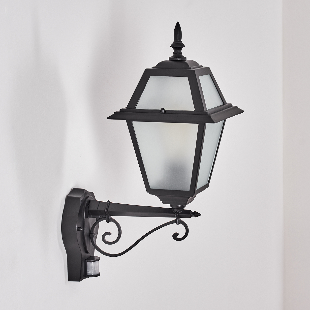 Eine schwarze Außenwandleuchte mit mattierten Glasscheiben. Sie ist an einer weißen Wand montiert und verfügt über einen Bewegungssensor.
