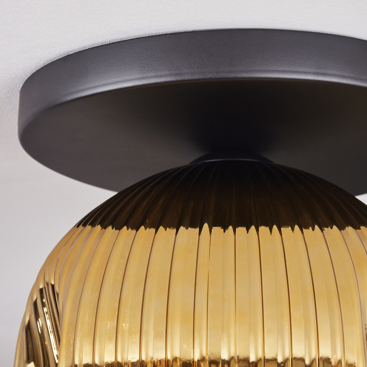 Nahaufnahme einer Deckenlampe. Sie hat einen schwarzen Sockel und einen gerippten, goldfarbenen Glasschirm, auf weißem Hintergrund.