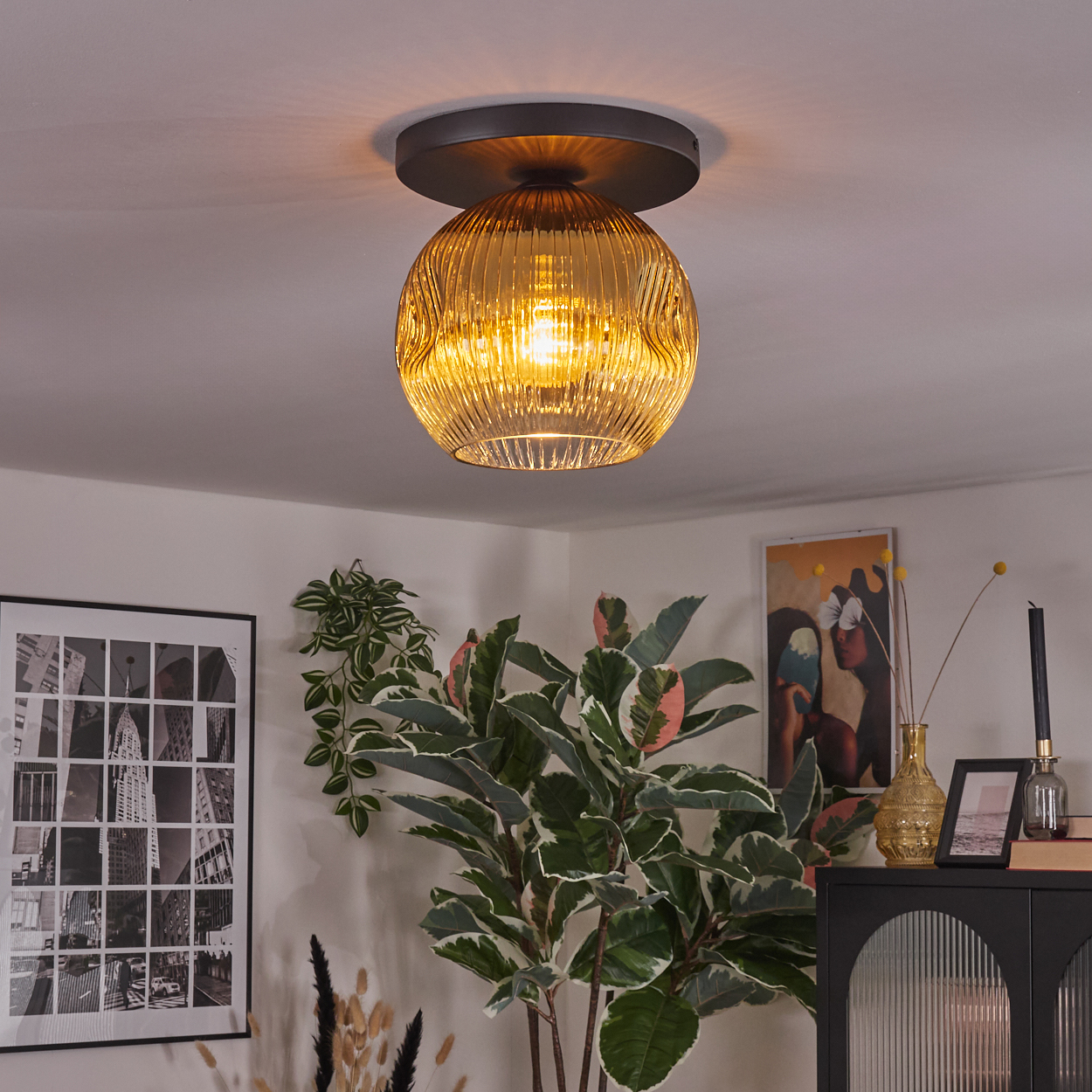 Dekorative Deckenlampe mit strukturiertem, bernsteinfarbenem Glasschirm. Die Umgebung umfasst Pflanzen, Kunstwerke und einen schwarzen Schrank.