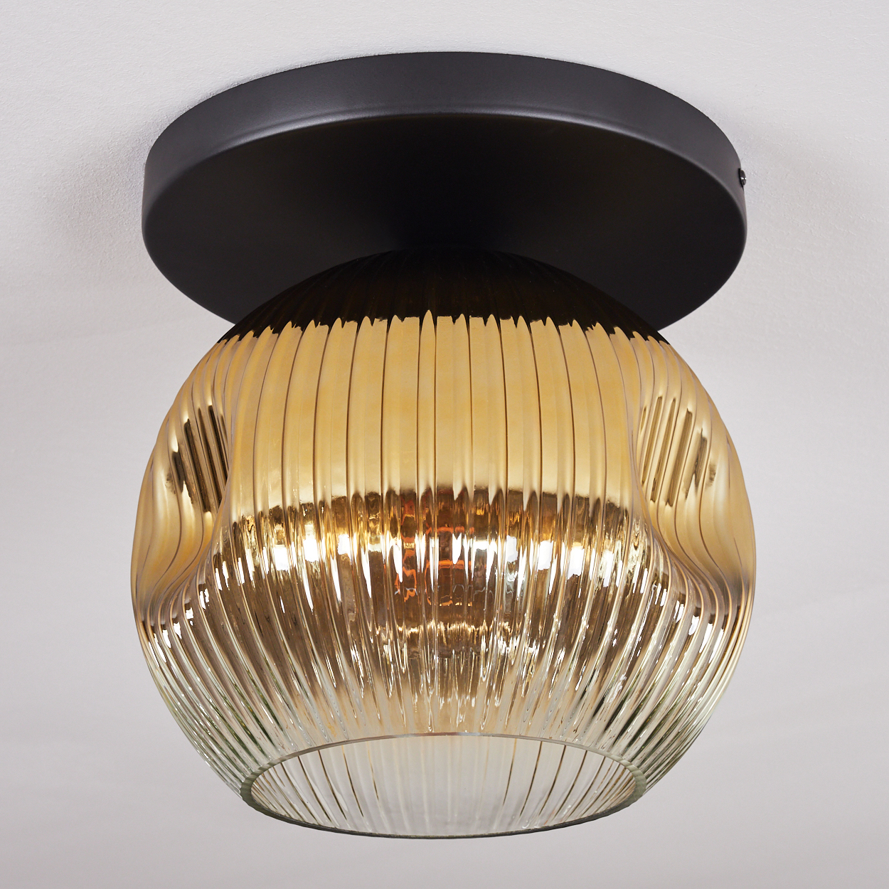 Eine moderne Deckenleuchte. Sie hat einen schwarzen Sockel und einen gerippten, goldfarbenen Glasschirm. Der Hintergrund ist eine weiße Decke.