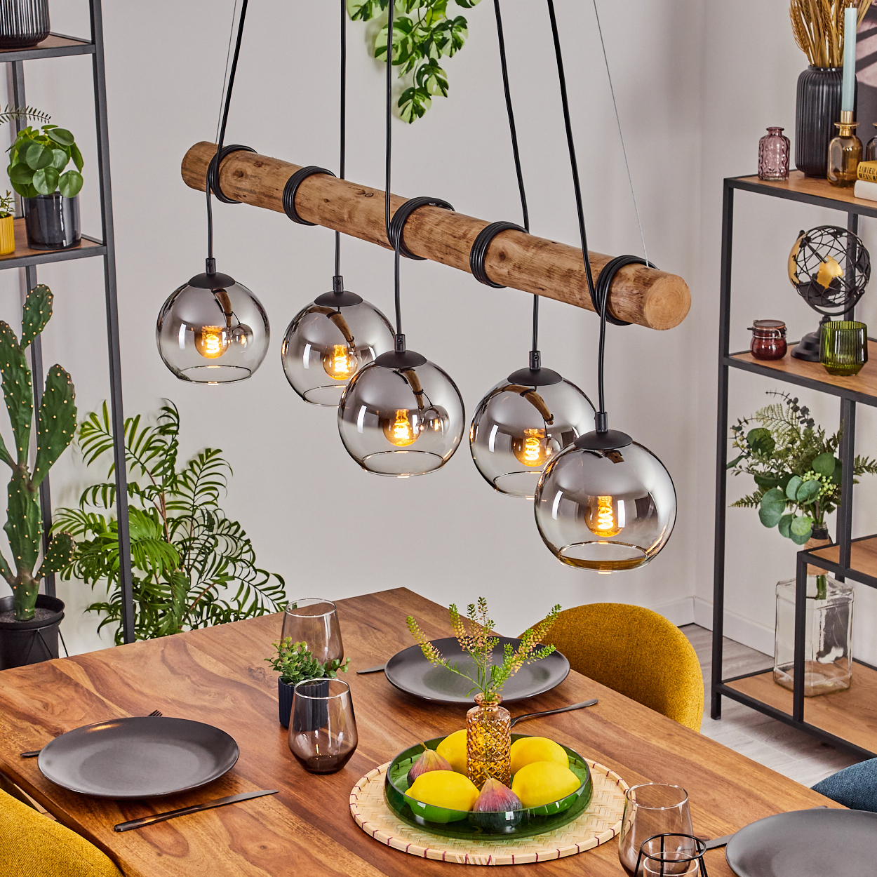 Ein Esszimmer mit Holztisch, Leuchte mit Glaskugeln und Obstteller. Regale mit Pflanzen im Hintergrund.